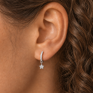 A close-up of a model's ear wearing a silver crescent-shaped huggie hoop earring with a dangling star charm set with clear stones.