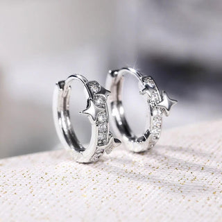 A pair of silver huggie hoop earrings decorated with star-shaped charms and small clear stones, displayed on a textured light beige surface.
