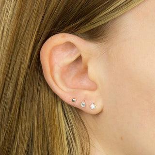 Close-up of a person's ear wearing three small silver stud earrings, including a round stud, a teardrop-shaped clear stone stud, and a star-shaped clear stone stud.