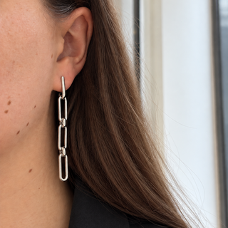 A close-up of a person's ear wearing a long, silver link drop earring. The person has long brown hair and is wearing a black top, with a softly blurred light-colored background.