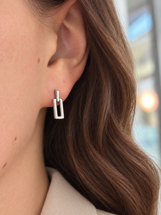 A close-up of a woman's ear wearing a small silver geometric drop earring with a rectangular frame design.