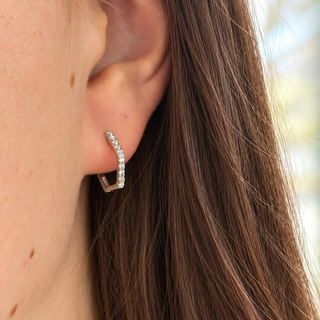 A close-up of a model's ear wearing a small silver hexagon-shaped hoop earring set with clear stones, with her brown hair flowing down.