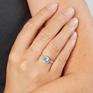 Hand wearing a ring with a blue gemstone, against a neutral background