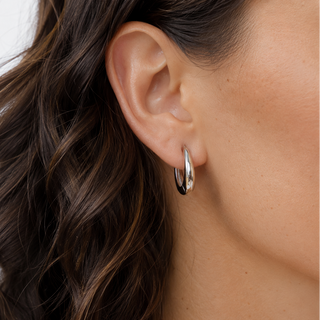 A close-up of a model's ear wearing a polished silver hoop earring, with her wavy brown hair and skin visible against a clean white background.