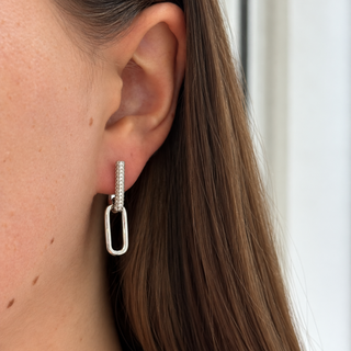 A close-up of a woman's ear wearing a silver drop earring with a small vertical pavé bar and a larger open oval drop, against her brown hair and skin.
