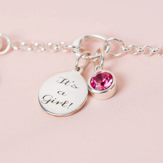 Silver bracelet with 'It's a Girl!' charm and pink gemstone on a pink background