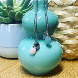 Two turquoise vases with necklaces hanging from them, placed on a wooden surface with a woven mat and potted plant in the background.