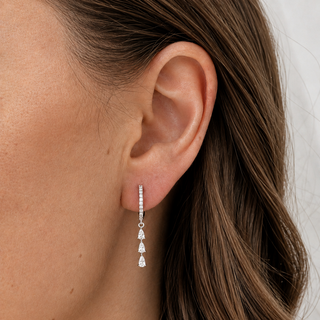A close-up of a model's ear wearing a delicate silver huggie hoop earring with three dangling teardrop-shaped clear stones.