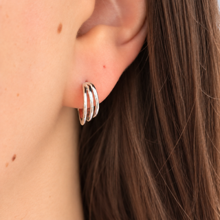 A close-up of a person's ear wearing a small silver hoop earring with three parallel curved bands, against dark brown hair and light skin.