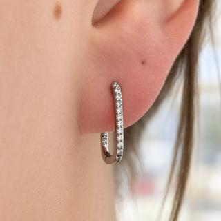 A close-up of a person's ear wearing a small silver huggie hoop earring set with clear stones. The inside of the hoop is stamped with '925', indicating sterling silver.