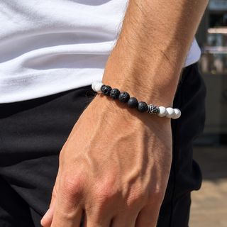 A close-up of a man's wrist wearing a beaded bracelet with black lava stones, white beads, and a small silver spacer, paired with a white t-shirt and black pants.