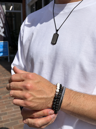 A close-up of a person wearing a black beaded bracelet and a black dog tag necklace outdoors. The person is wearing a white t-shirt, and the background shows a brick pavement and storefront.