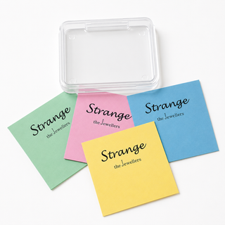Four square pastel sticky notes in green, pink, yellow, and blue, each printed with the text 'Strange the Jewellers' in black script, arranged on a white background with a clear plastic storage box behind them.