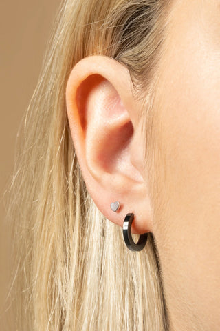 Close-up of an ear wearing a small silver heart earring and a black hoop earring on a beige background.