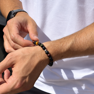 A close-up of a man's hands adjusting a beaded bracelet on his wrist. The bracelet features dark brown, black, and polished gold-toned beads.