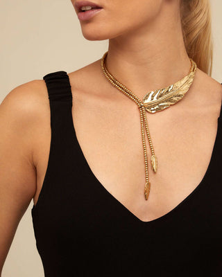 A close-up of a model wearing a gold-toned necklace with a large feather-shaped centerpiece and dangling bead strands, paired with a black top against a warm beige background.