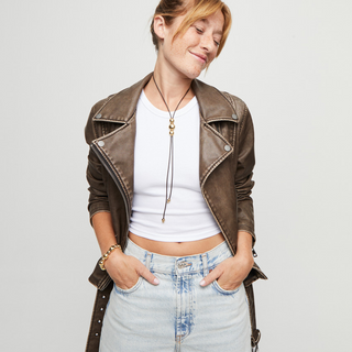 Woman wearing a brown leather jacket, white top, and light blue jeans on a plain background