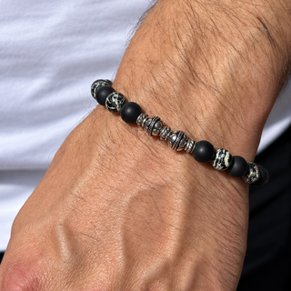 A close-up of a man's wrist wearing a beaded bracelet with matte black beads, silver metallic spacer beads, and skull-shaped beads.