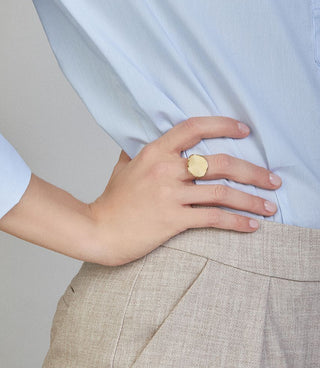 Person wearing a light blue shirt and beige pants with a gold ring on a neutral background