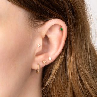 Close-up of an ear with multiple earrings including a gold hoop, heart-shaped earring, and green gemstone.